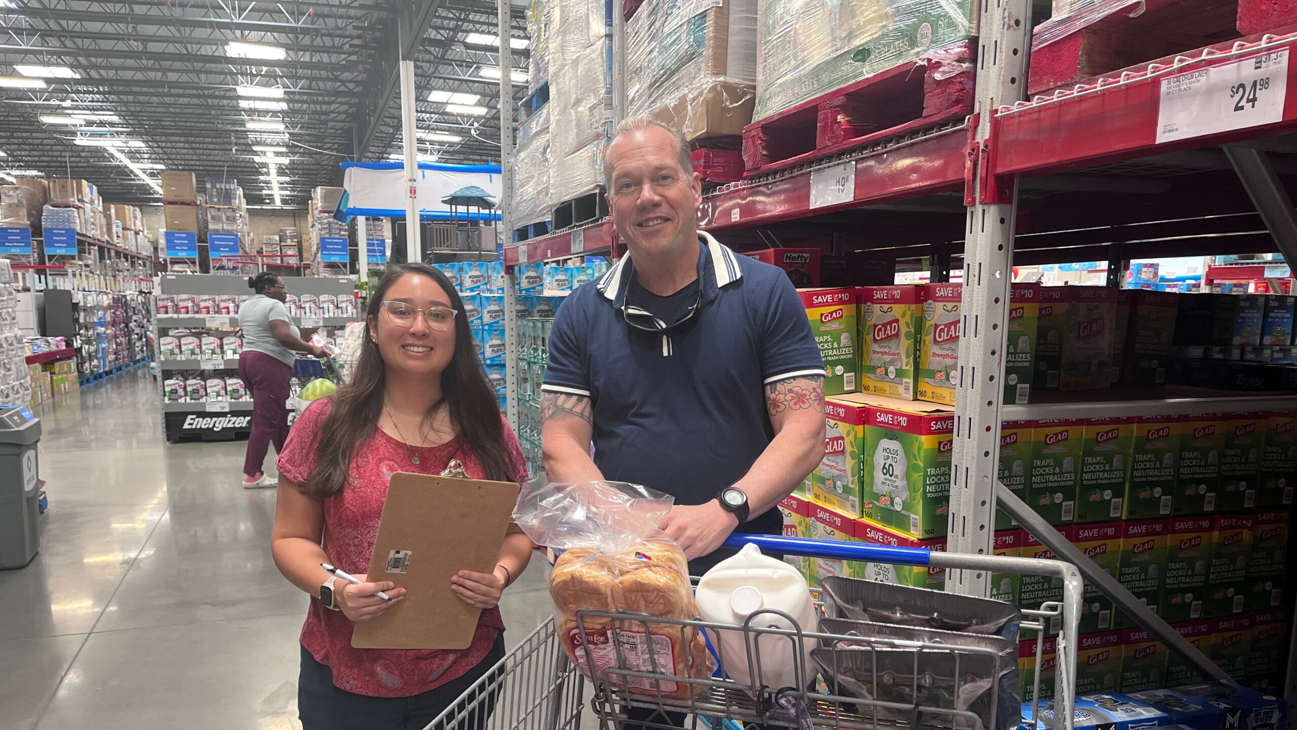 Two people posing for a photo at a warehouse supermarket with a shopping cart full of groceries